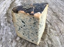 A picture of a wedge of Valdeon Picos Blue PGI Cheese. The cheese is covered in sycamore leaves and is positioned on a rustic wooden table