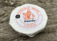 A picture of a wrapped wheel of Waterloo Cheese. It has been placed on a rustic wooden table.
