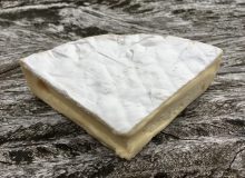A picture of a quarter wheel of West Country Brie Cheese. It is positioned on a rustic, wooden table.